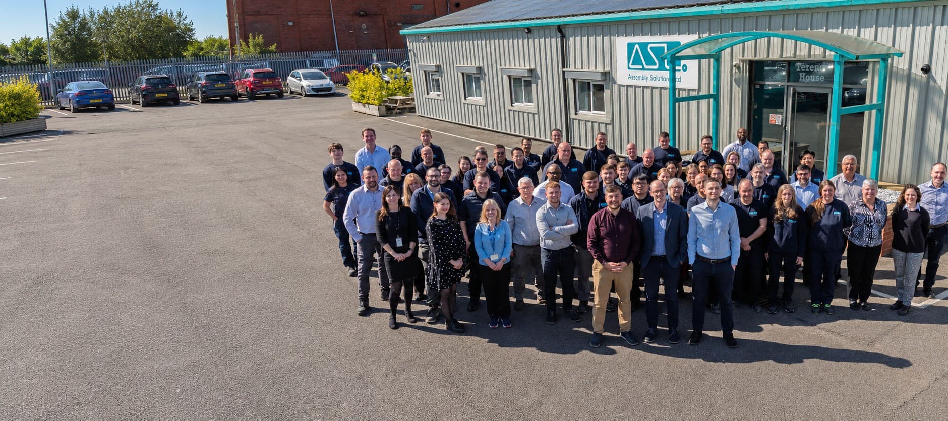 The ASL team, around eighty people in navy company shirts and office wear, gathered in the sunshine outside Terence House, ASL's factory in Bolton, under the ASL Assembly Solutions sign
