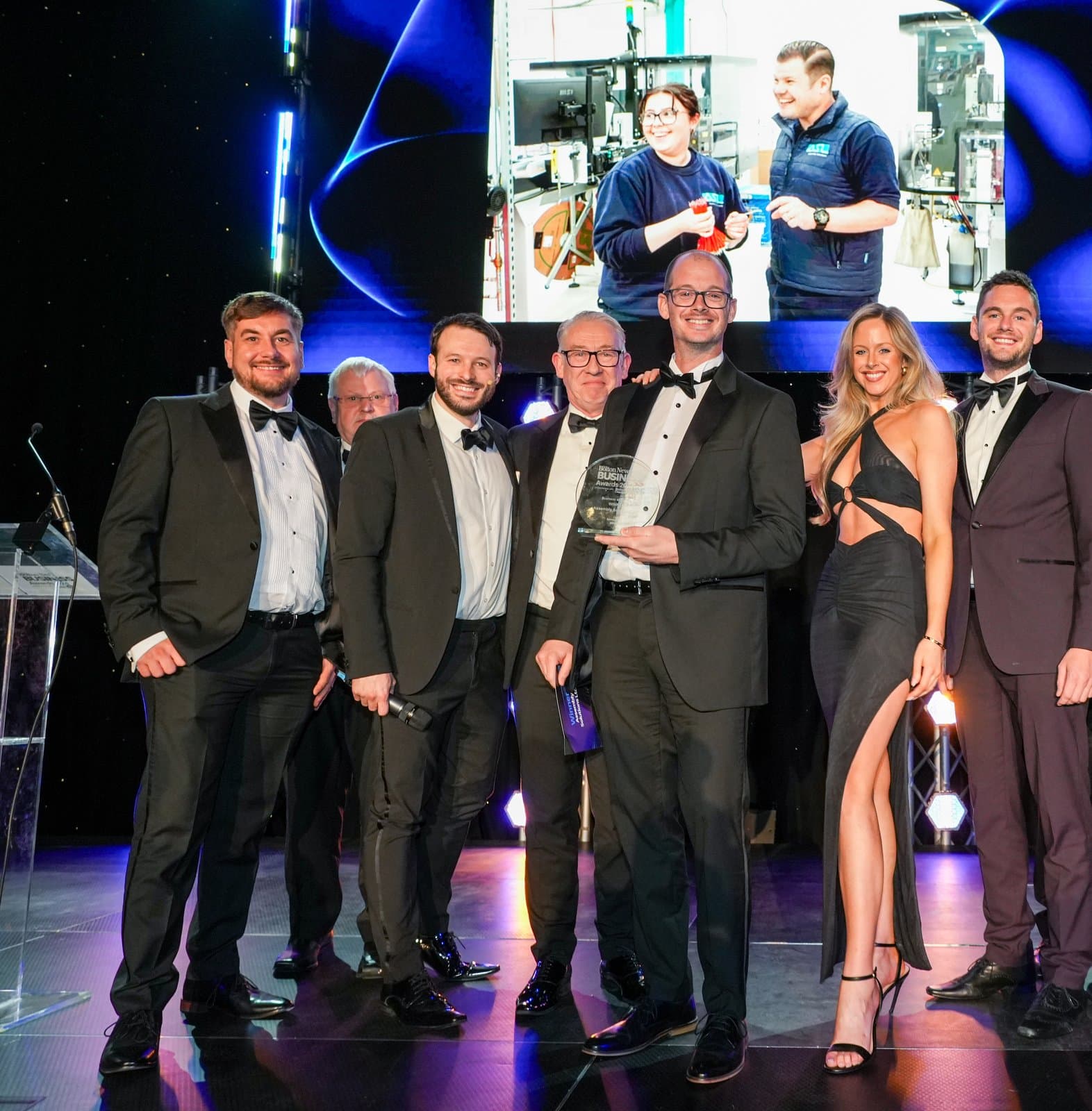 The ASL team on stage at the Bolton News Business Awards 2024, in evening dress, holding the glass Business of the Year trophy, with a photo of two ASL colleagues at work on the screen behind them