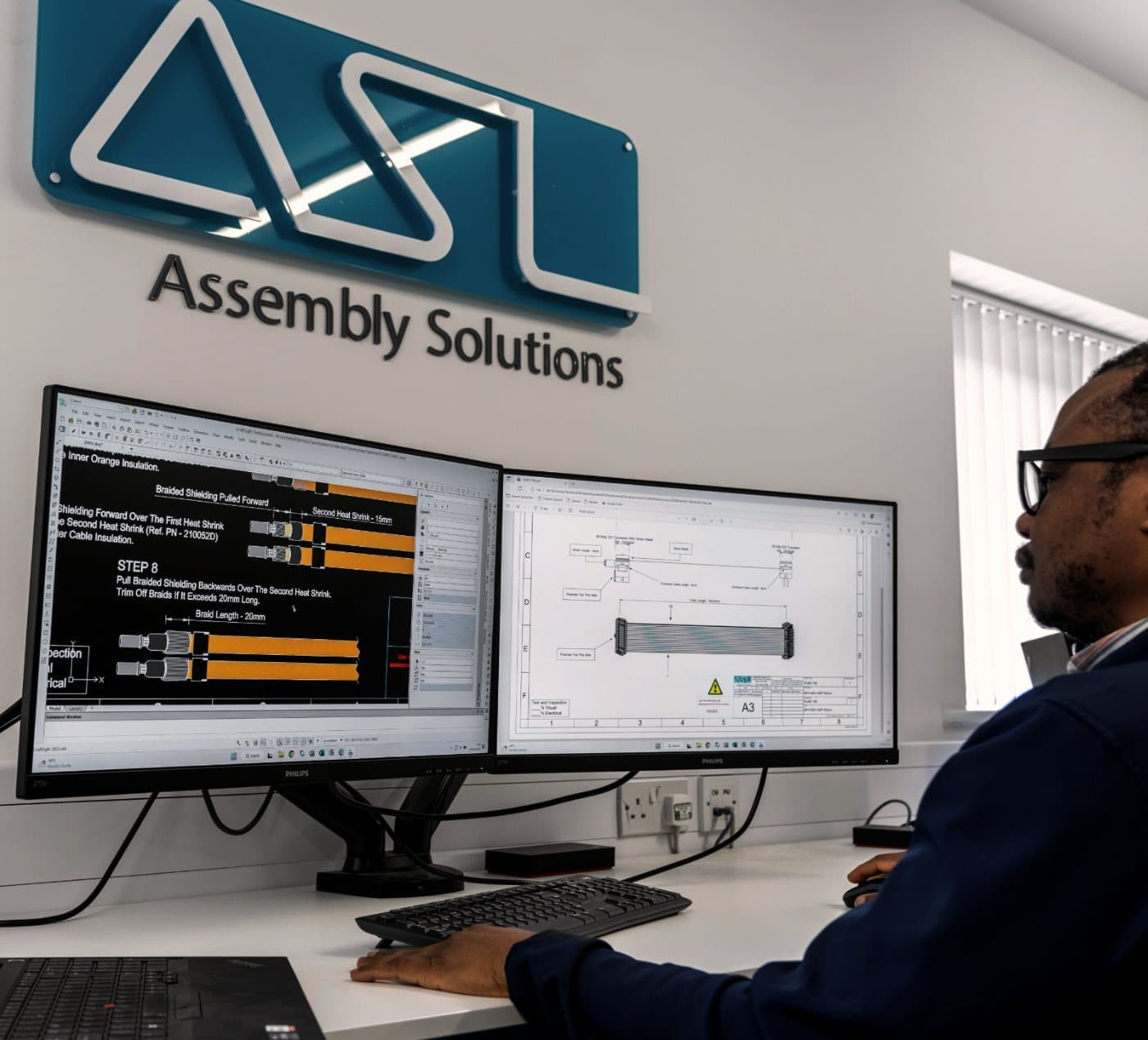 An ASL engineer at a two-screen workstation beneath the Assembly Solutions sign, preparing step-by-step cable assembly instructions beside an assembly drawing with an ASL title block